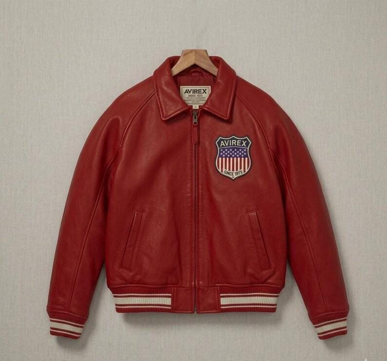 May include: A bright red leather jacket featuring a classic collar, a full-length front zipper, and ribbed cuffs and hem with white stripes. A shield-shaped patch with the text AVIREX and a flag design is on the chest, with an AVIREX label at the neck.