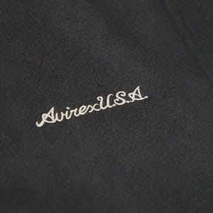 Op de afbeelding: Een zwart stoffen oppervlak met de wit geborduurde tekst AvirexU.S.A. in een cursief lettertype. Het borduurwerk is diagonaal geplaatst op het donkere, getextureerde materiaal en toont duidelijke letters en stikseldetails.