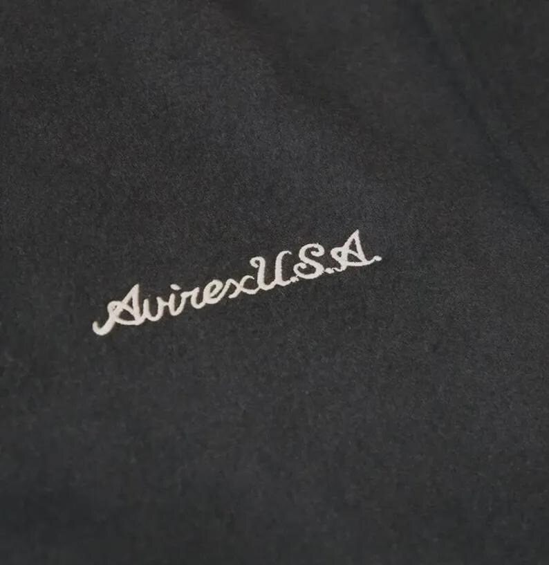 Op de afbeelding: Een zwart stoffen oppervlak met de wit geborduurde tekst AvirexU.S.A. in een cursief lettertype. Het borduurwerk is diagonaal geplaatst op het donkere, getextureerde materiaal en toont duidelijke letters en stikseldetails.