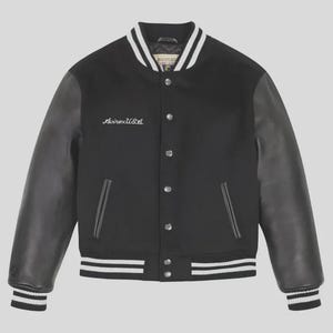 Op de afbeelding: Een zwart varsity-jack met zwarte leren mouwen en een zwarte body van wolmix. Het jack heeft een drukknoopsluiting, twee zijzakken en wit gestreepte geribbelde boorden bij de kraag, mouwen en zoom. AvirexUSA is in het wit op de borst geborduurd.