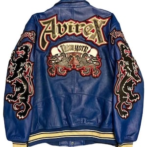 May include: Blue leather jacket with embroidered patches. The back features the word "Avirex" in large letters, a "New York" banner, and two lion emblems. The sleeves have additional lion designs. The jacket has a yellow and white striped trim.