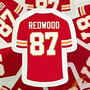 Red football jersey sticker with “REDWOOD 87,” inspired by Taylor Swift’s Life of a Showgirl “wood” song and Travis Kelce. Features white and yellow sleeve stripes and a glossy laminated finish.