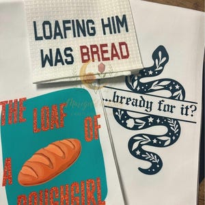 May include: White kitchen towels with humorous text and designs. One towel reads "LOAFING HIM WAS BREAD" in navy and red. Another features a bread loaf graphic with "THE LOAF OF A DOUGHGIRL" in orange glitter. A third towel has a snake design.