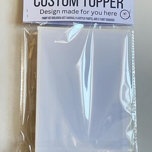 Op de afbeelding: Een doorzichtige plastic verpakking met een witte kaart met de tekst "CUSTOM TOPPER" en "Design made for you here" gedrukt op een wit etiket. Het pakket bevat ook een schilderset.