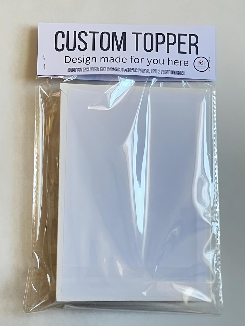 Op de afbeelding: Een doorzichtige plastic verpakking met een witte kaart met de tekst "CUSTOM TOPPER" en "Design made for you here" gedrukt op een wit etiket. Het pakket bevat ook een schilderset.