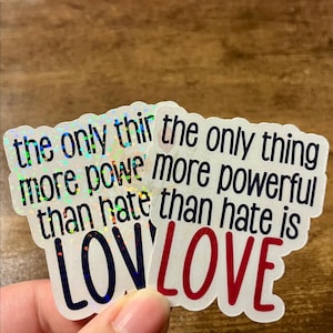 May include: Two stickers with the text "the only thing more powerful than hate is LOVE". The stickers have a holographic, iridescent finish, with black and red lettering on a white background.