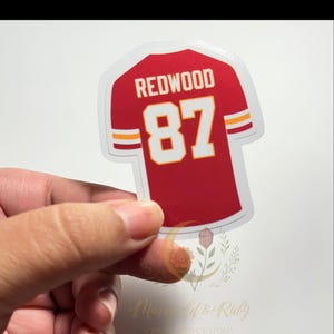 hand holding Swiftie sticker inspired by Taylor Swift’s Life of a Showgirl “wood” song and Travis Kelce. Red football jersey design with “REDWOOD 87,” glossy laminate, and white-yellow-white sleeve stripes.