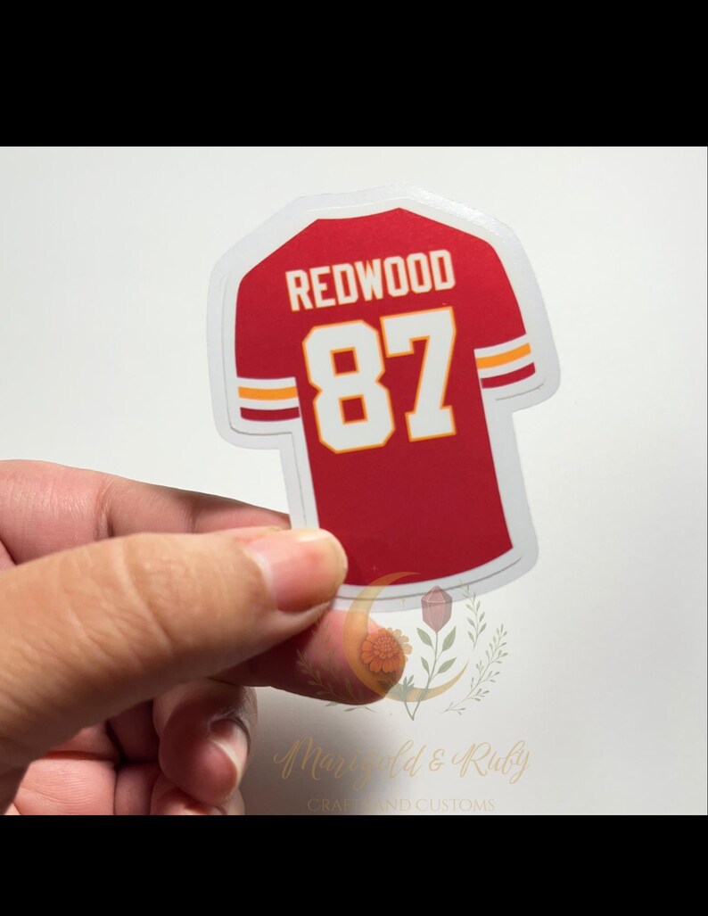 hand holding Swiftie sticker inspired by Taylor Swift’s Life of a Showgirl “wood” song and Travis Kelce. Red football jersey design with “REDWOOD 87,” glossy laminate, and white-yellow-white sleeve stripes.