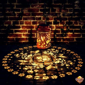 May include: A solar-powered lantern with a floral and hummingbird design, casting a decorative light pattern on the ground. The lantern is a reddish-brown color and hangs from a metal handle. The background is a brick wall.