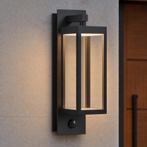 May include: A rectangular outdoor wall lantern featuring a matte black metal frame with clear glass panels. The fixture includes an integrated light source at the top and a small circular motion sensor located on the lower mounting plate section.