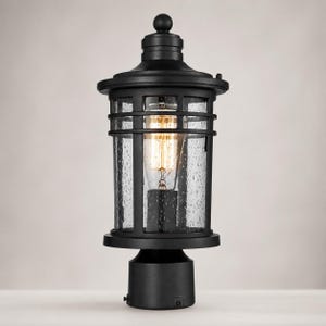 May include: A matte black outdoor post light fixture featuring a cylindrical frame with a decorative top finial. The lantern includes clear seeded glass panels that enclose a single vintage-style Edison bulb, providing a warm glow.