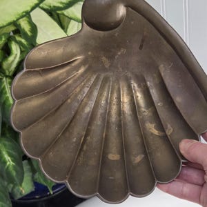 May include: A vintage brass seashell-shaped dish with a scalloped edge. The dish has a textured surface and a curved handle. The brass has a warm, aged patina, with some areas showing wear. The dish is held by a hand.