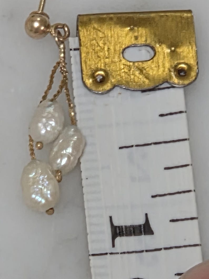 May include: A gold-toned earring with three white, irregularly shaped pearls dangling from a delicate chain. The earring is next to a measuring tape, showing the size of the pearls.
