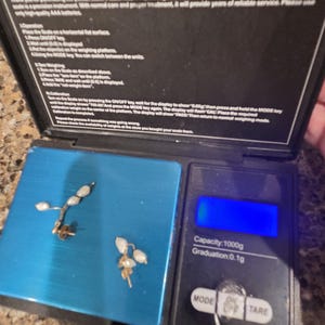 May include: Two gold-colored earrings with small, white, pearl-like beads are displayed on a blue digital scale. The scale's display is blank. The scale has a capacity of 1000g and a graduation of 0.1g.