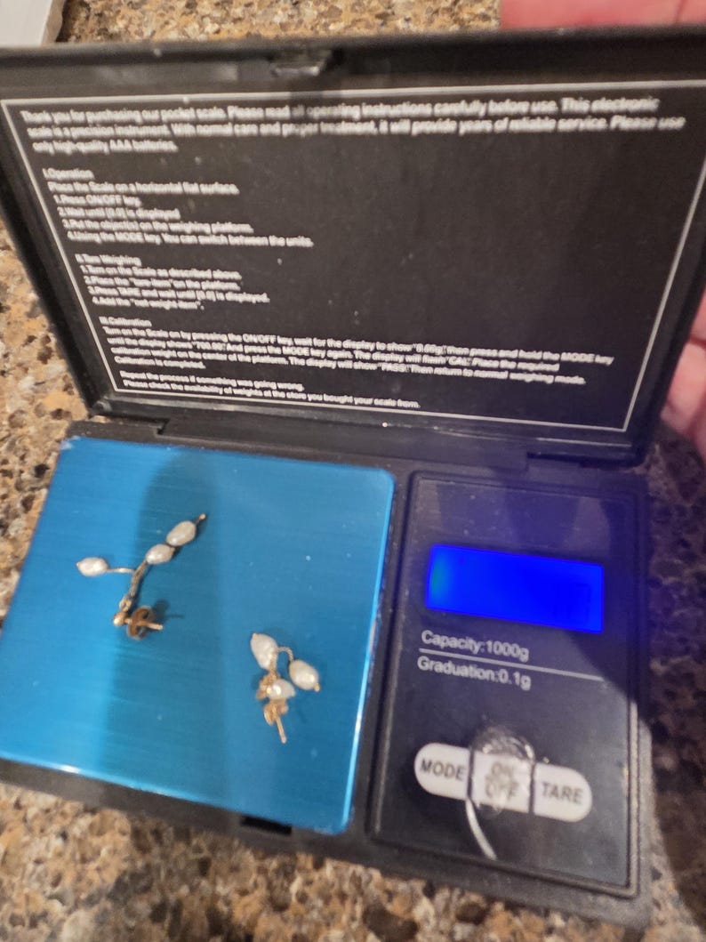 May include: Two gold-colored earrings with small, white, pearl-like beads are displayed on a blue digital scale. The scale's display is blank. The scale has a capacity of 1000g and a graduation of 0.1g.