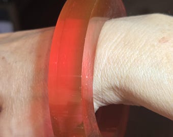 May include: A translucent, orange-red bangle bracelet. The thick, rounded bracelet is worn on a wrist, showcasing its smooth, polished surface. The color transitions from a lighter orange to a deeper red, creating a gradient effect. The bangle is a bold, statement accessory.