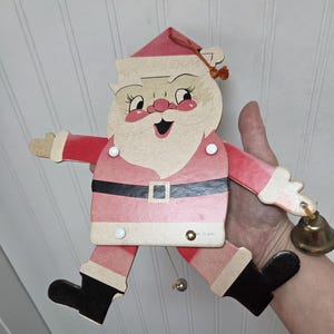 May include: A vintage Santa Claus decoration with a red and pink color scheme. The Santa figure has a smiling face, black boots, and a black belt. A small gold bell hangs from one arm, and a silver bell hangs from the bottom.