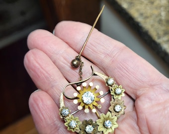 May include: A gold-toned brooch with a floral design and clear rhinestones. The brooch has a pin and a dangling chain with a small ball at the end.
