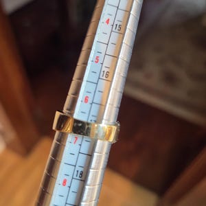 May include: A gold ring being sized on a metal ring sizer. The ring sizer has markings from 3 to 9, with numbers 14, 15, 16, 18, and 19 visible.