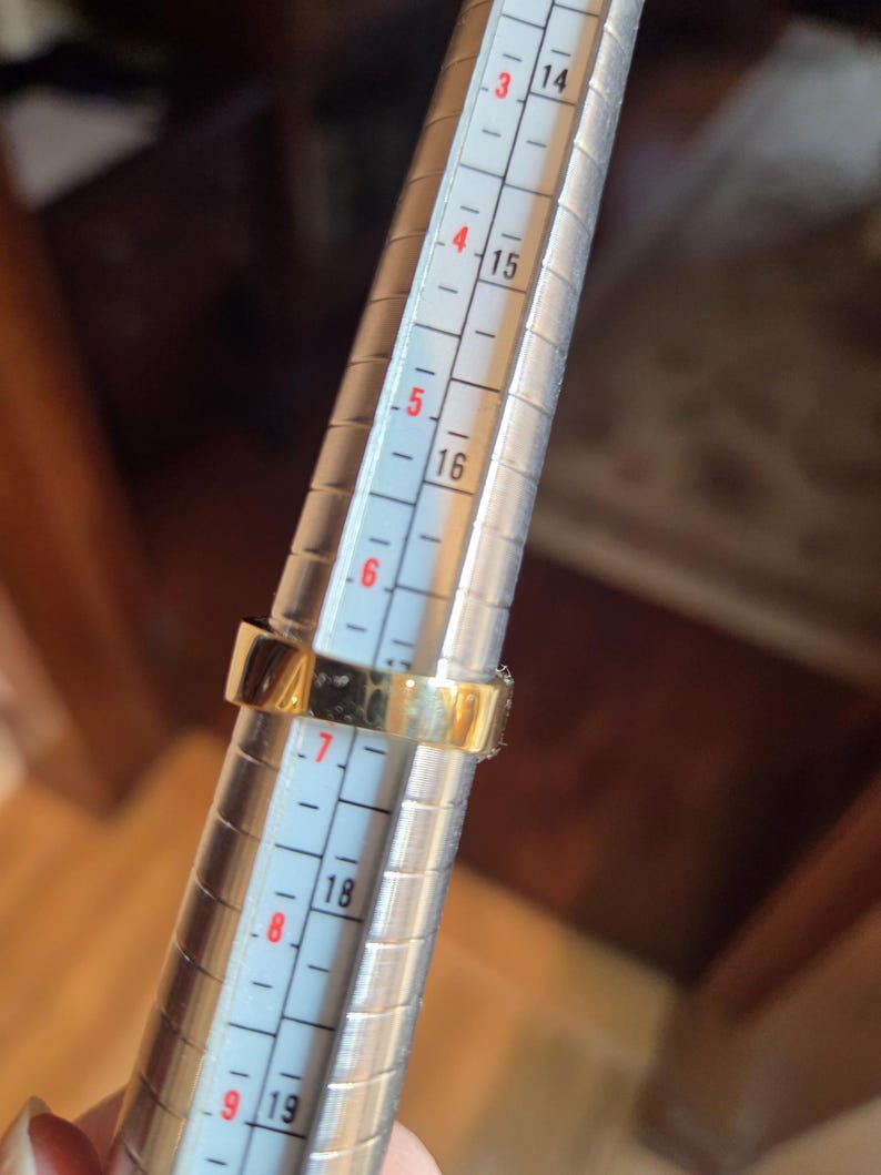 May include: A gold ring being sized on a metal ring sizer. The ring sizer has markings from 3 to 9, with numbers 14, 15, 16, 18, and 19 visible.