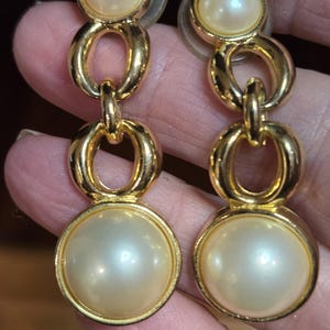 May include: A pair of gold-toned drop earrings featuring faux pearls. Each earring has a large, round pearl at the bottom, a smaller pearl at the top, and two gold chain-link style connectors. The earrings are designed to be worn with pierced ears.