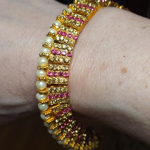 May include: A gold bracelet with pink and white stones. The bracelet is made of small, square-shaped pieces that are linked together.