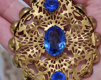 May include: A gold filigree pendant necklace with three blue glass stones. The pendant is in the shape of a flower with a delicate design.