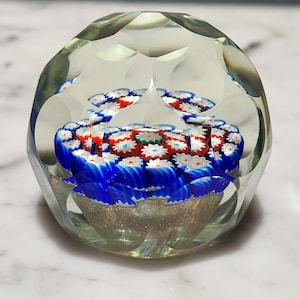 May include: A clear glass paperweight with a multi-colored, flower-like design inside. The paperweight is faceted and has a round shape.
