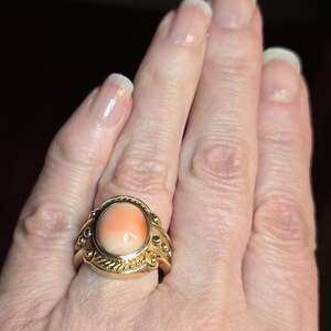 May include: A gold ring with a large oval-shaped coral stone set in the center. The ring has a detailed design with small gold beads and a rope-like pattern around the stone.
