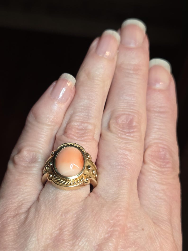May include: A gold ring with a large oval-shaped coral stone set in the center. The ring has a detailed design with small gold beads and a rope-like pattern around the stone.