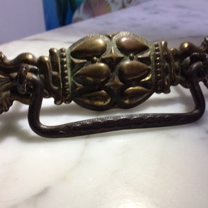 May include: A vintage brass drawer pull with a decorative, ornate design. The pull has a curved, metal handle.