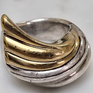 May include: A silver and gold ring with a swirling design. The ring is made of two metals, with the gold metal forming a wave-like pattern on top of the silver metal.