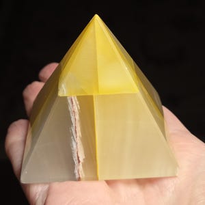 May include: A translucent, yellow and white pyramid with a pointed top. The pyramid is held in a hand against a black background. The pyramid has a natural, layered appearance with a reddish-brown streak.
