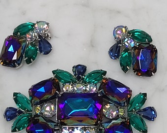 May include: A set of iridescent blue and green rhinestone jewelry featuring a large brooch and a pair of earrings. The brooch is a cluster of faceted rhinestones in shades of blue and green, with a large rectangular center stone. The earrings are smaller versions of the brooch, with a single large rectangular stone.