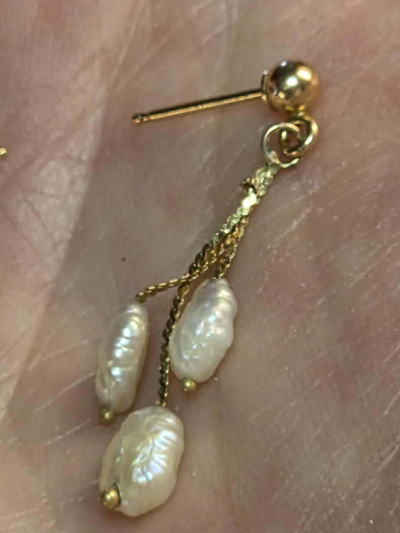 May include: A gold-colored earring with three dangling, irregularly shaped white pearls. The earring has a post and a small gold ball at the top. The pearls are attached by a delicate gold chain.