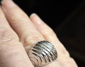 May include: A silver ring with a ridged, dome-shaped design. The ring is worn on a finger, and the ridges create a textured surface. The ring's metallic sheen reflects light, highlighting the detailed pattern. The ring is a piece of jewelry.
