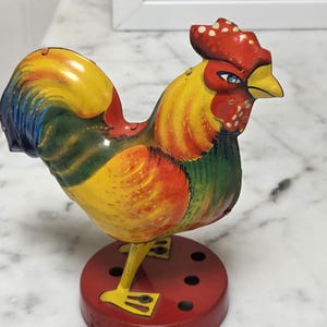 May include: A colorful, vintage metal rooster figurine. The rooster features a red comb, yellow beak, and a body painted with yellow, green, and blue hues. It stands on a red circular base with several holes.