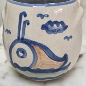 May include: A ceramic mug with a whimsical design. The off-white mug features a blue rim and is decorated with a blue and tan sea creature, blue clouds, and small blue birds. The mug has a rounded shape and two small handles.