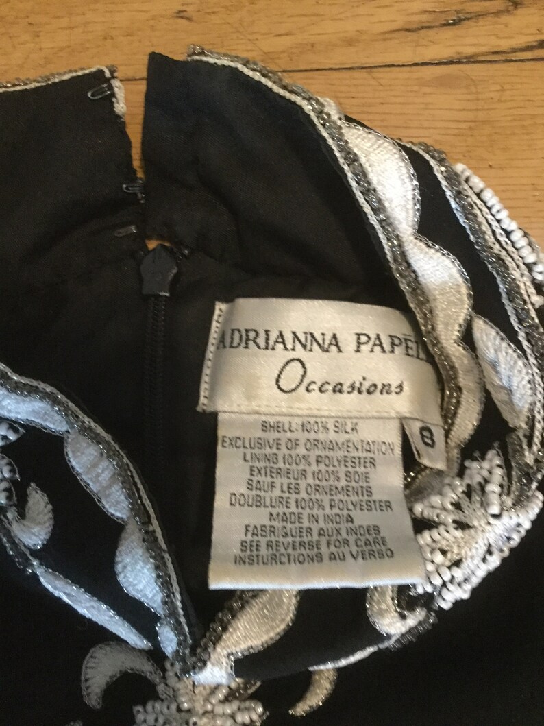 Puede incluir: Vestido negro de seda con bordados blancos y plateados. El vestido tiene una etiqueta que dice "Adrianna Papell Occasions" e incluye instrucciones de cuidado. La etiqueta de talla indica la talla 8.