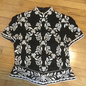 Puede incluir: Una blusa negra de manga corta con un patr&oacute;n de bordado floral blanco.