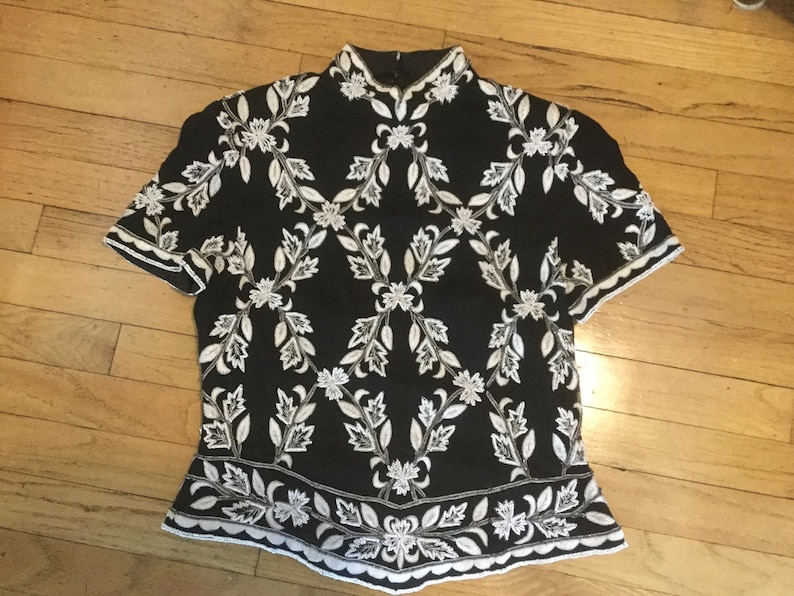 Puede incluir: Una blusa negra de manga corta con un patr&oacute;n de bordado floral blanco.