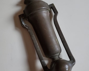 May include: A vintage brass door knocker with a decorative design. The knocker is shaped like a vase with a rounded top and a ring handle.