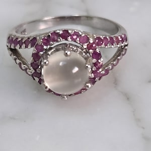 May include: A silver ring featuring a large, oval, milky-white gemstone at the center, surrounded by a halo of small, round, pink gemstones. The ring band is also adorned with pink gemstones.