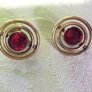 May include: A pair of vintage gold-toned earrings with a circular design. Each earring features a large, round, deep red gemstone set in the center, surrounded by concentric circles. The earrings are displayed on a light green textured surface.