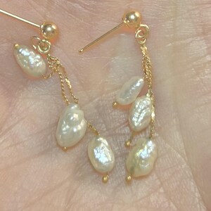 May include: Gold-colored dangle earrings featuring teardrop-shaped white pearls. The earrings have a post-and-ball closure, with delicate gold chains suspending the pearls. The pearls have a lustrous, irregular surface.