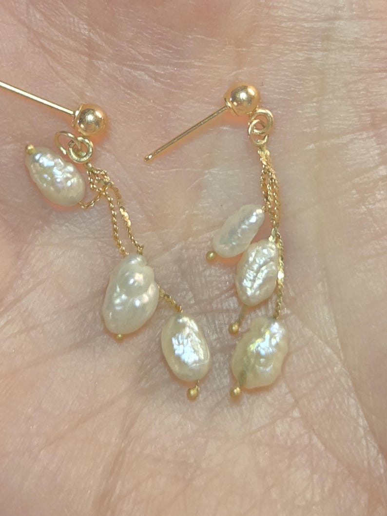 May include: Gold-colored dangle earrings featuring teardrop-shaped white pearls. The earrings have a post-and-ball closure, with delicate gold chains suspending the pearls. The pearls have a lustrous, irregular surface.