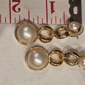 May include: A pair of gold-toned drop earrings featuring faux pearls. Each earring has a large, round pearl, connected by gold chain links to a smaller pearl. The earrings are approximately 1 inch in length.