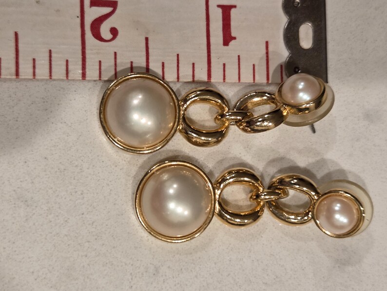 May include: A pair of gold-toned drop earrings featuring faux pearls. Each earring has a large, round pearl, connected by gold chain links to a smaller pearl. The earrings are approximately 1 inch in length.