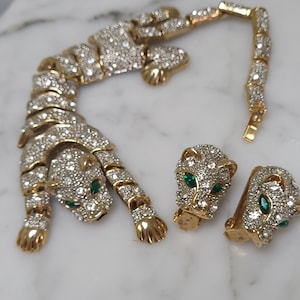 May include: A gold tone, rhinestone-encrusted, crouching panther necklace and matching earrings with green emerald eyes.