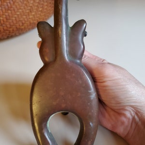 May include: A bronze cat figurine with a curled tail, standing on its hind legs. The cat is facing away from the camera, with its back and tail visible. The figurine has a rustic, aged appearance.